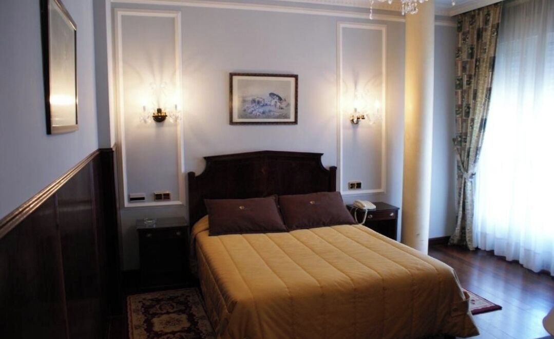 Une chambre d’hôtel avec un grand lit, deux tables de chevet, une commode, un tableau encadré au mur et un lustre.