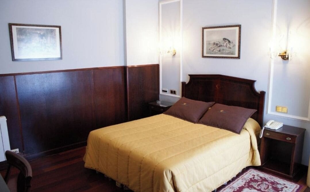 A hotel room with a large bed, wooden headboard, two bedside tables, a nightstand, a telephone, and two wall-mounted light fixtures.