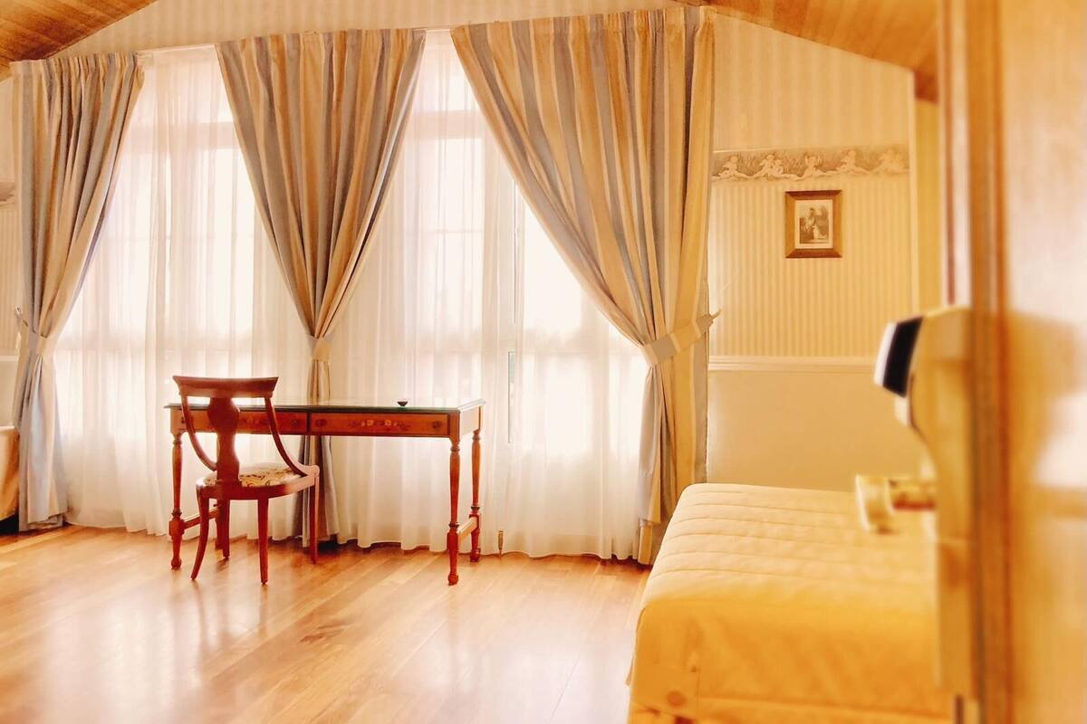 Una habitación con techo de madera, un escritorio de madera con una silla, una cama y ventanas amplias con cortinas.