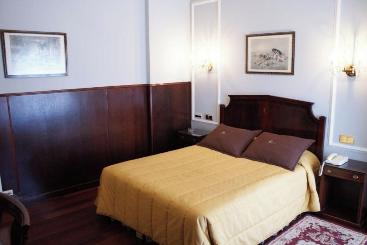 Habitación de hotel con una cama grande, cabecera de madera, dos mesitas de noche, un teléfono y dos lámparas fijadas a la pared.
