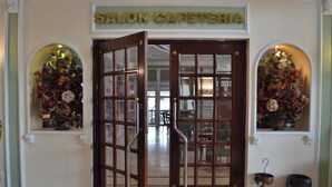 Un salon-cafétéria doté d’une porte en bois et de panneaux de verre, avec deux compositions florales de chaque côté, et une poubelle avec un parapluie à proximité.