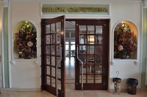 Cafetería con puerta de madera y paneles de vidrio, dos arreglos florales a cada lado y un cubo de basura con paraguas.