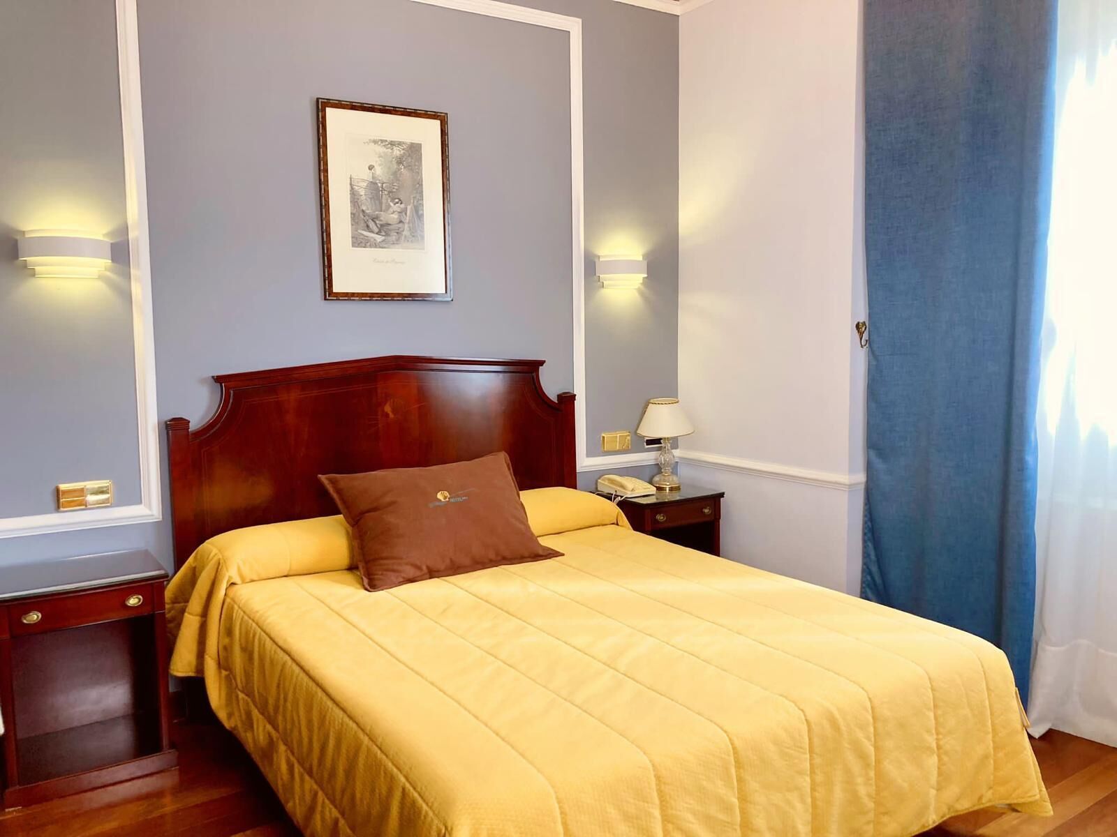 A neatly arranged hotel room with a wooden headboard, a yellow bedspread, a brown pillow, a framed picture on the wall, and a nightstand with a lamp.