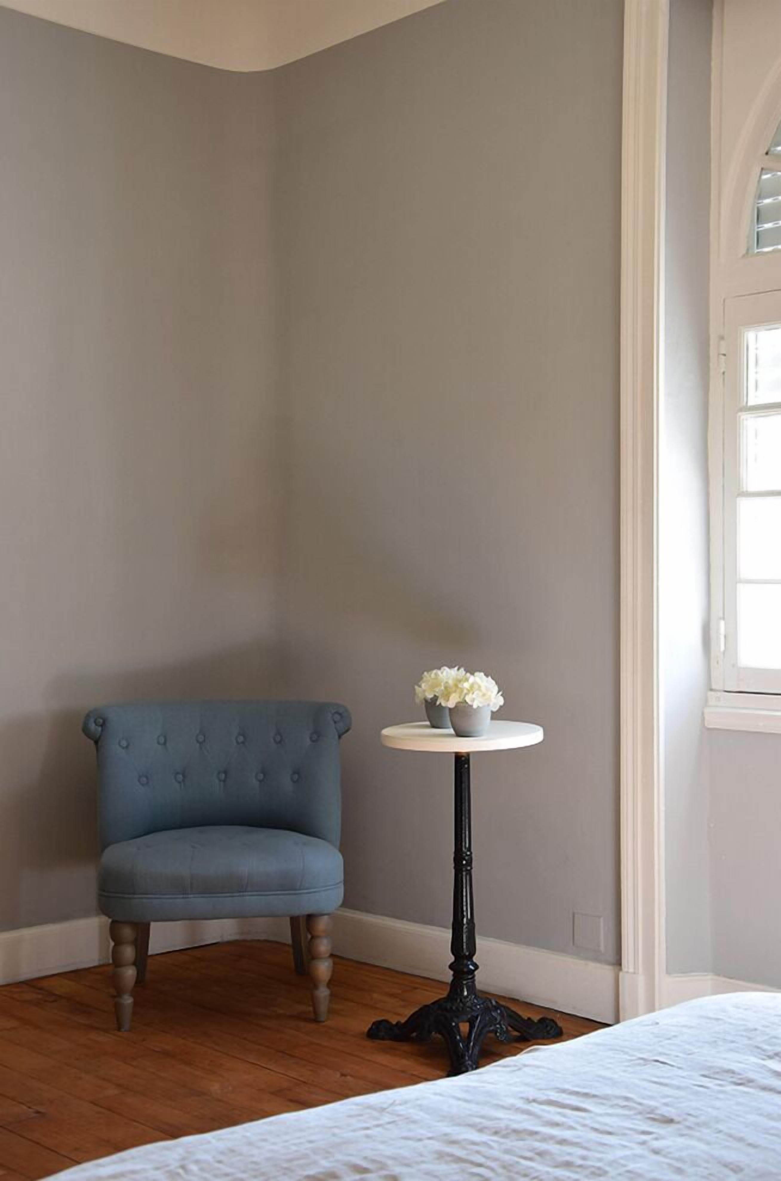 Una habitación de esquina con una butaca tapizada de azul, una mesita redonda con un jarrón de flores y una ventana con persianas.