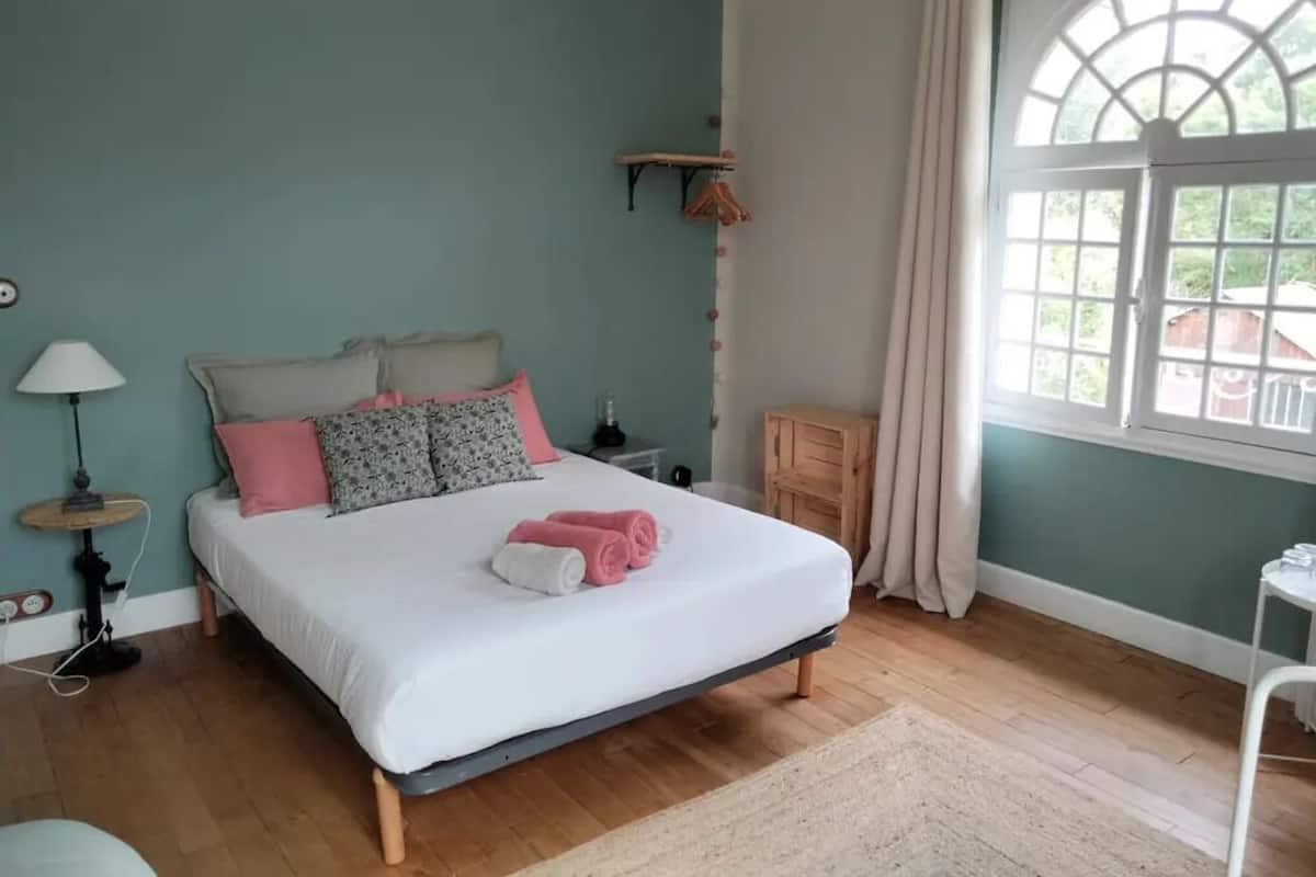 A bedroom with a bed, a nightstand, a lamp, a window with curtains, and a wooden floor.
