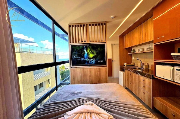 Ein modernes Hotelzimmer mit großem Fenster, Flachbildfernseher, Holzschränken und einer Küchenzeile.