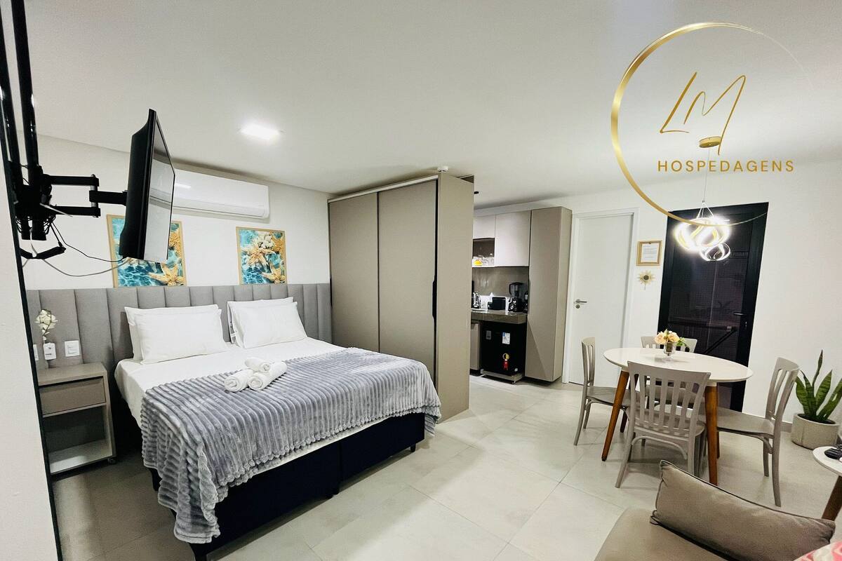 A modern hotel room with a bed, a desk, a dining area, and a kitchenette.