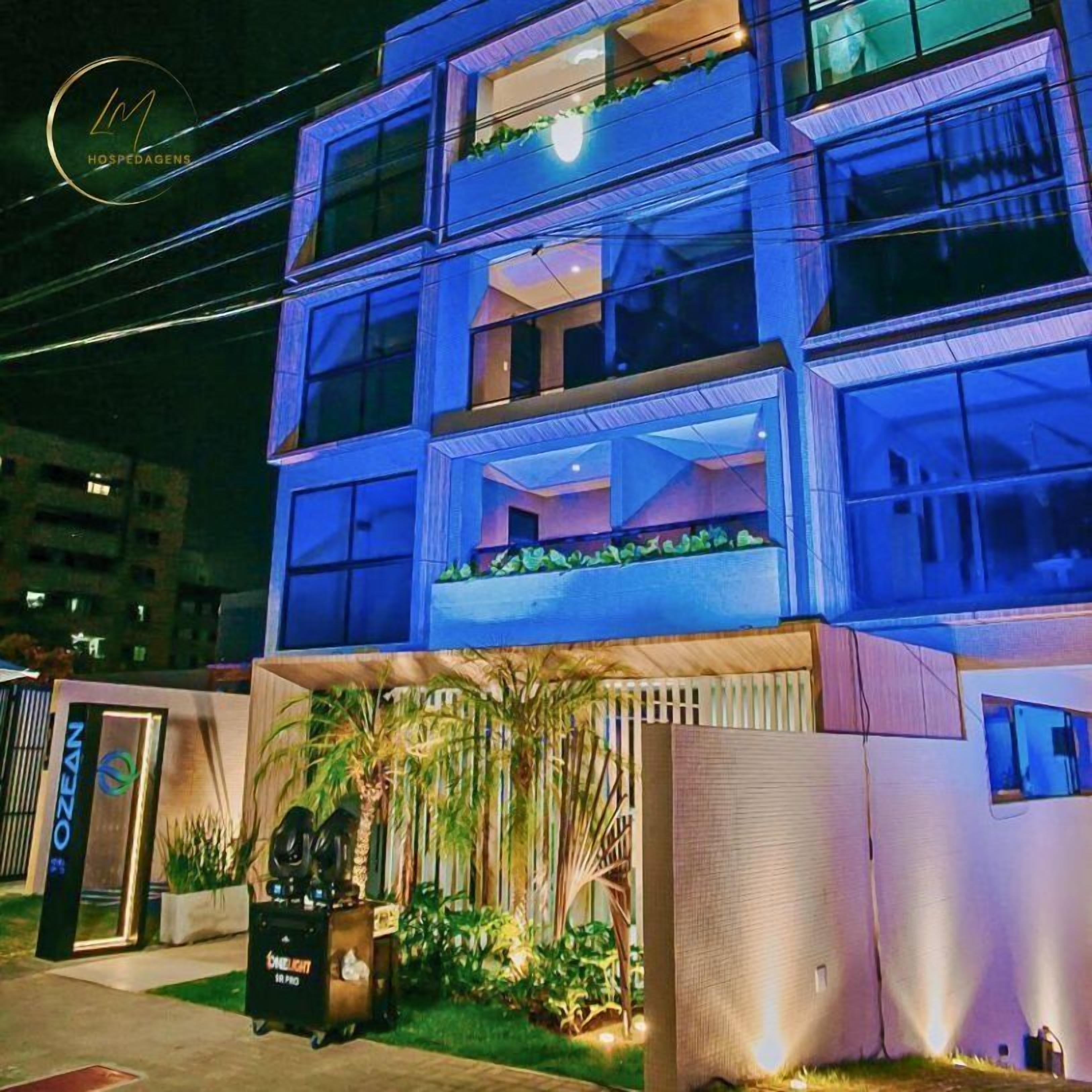 Un hotel de varios pisos con balcones, entrada protegida por un portón y plantas ornamentales.