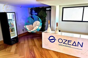 A reception area with a vending machine, a hanging chair, and a reception desk with the logo 'OZEAN HOME & SERVICE'. - LM OZEAN (Joao Pessoa)