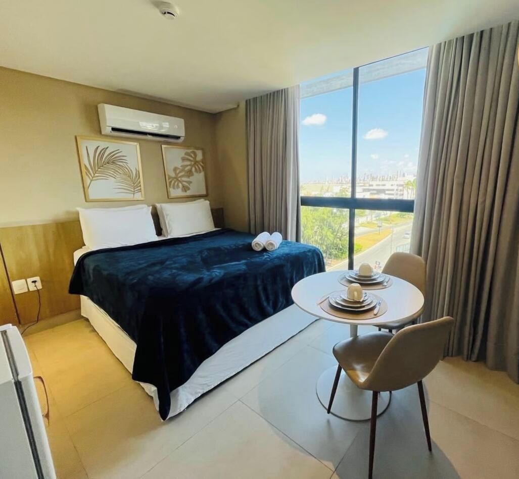 Habitació d'hotel amb llit, taula i una cadira, i una finestra gran que ofereix vista a la ciutat.