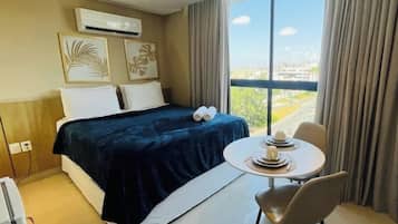 A hotel room with a bed, a table with a chair, and a large window offering a city view.