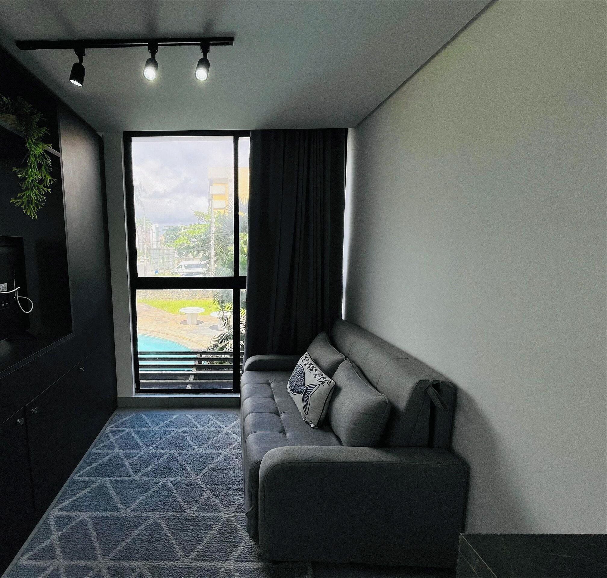 Una sala moderna con un sofá gris, un tapete estampado y un ventanal que ofrece vistas a una piscina y edificios.