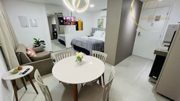 Una habitación de hotel moderna con zona de comedor, una cama con manta gris, televisión y una lámpara decorativa.
