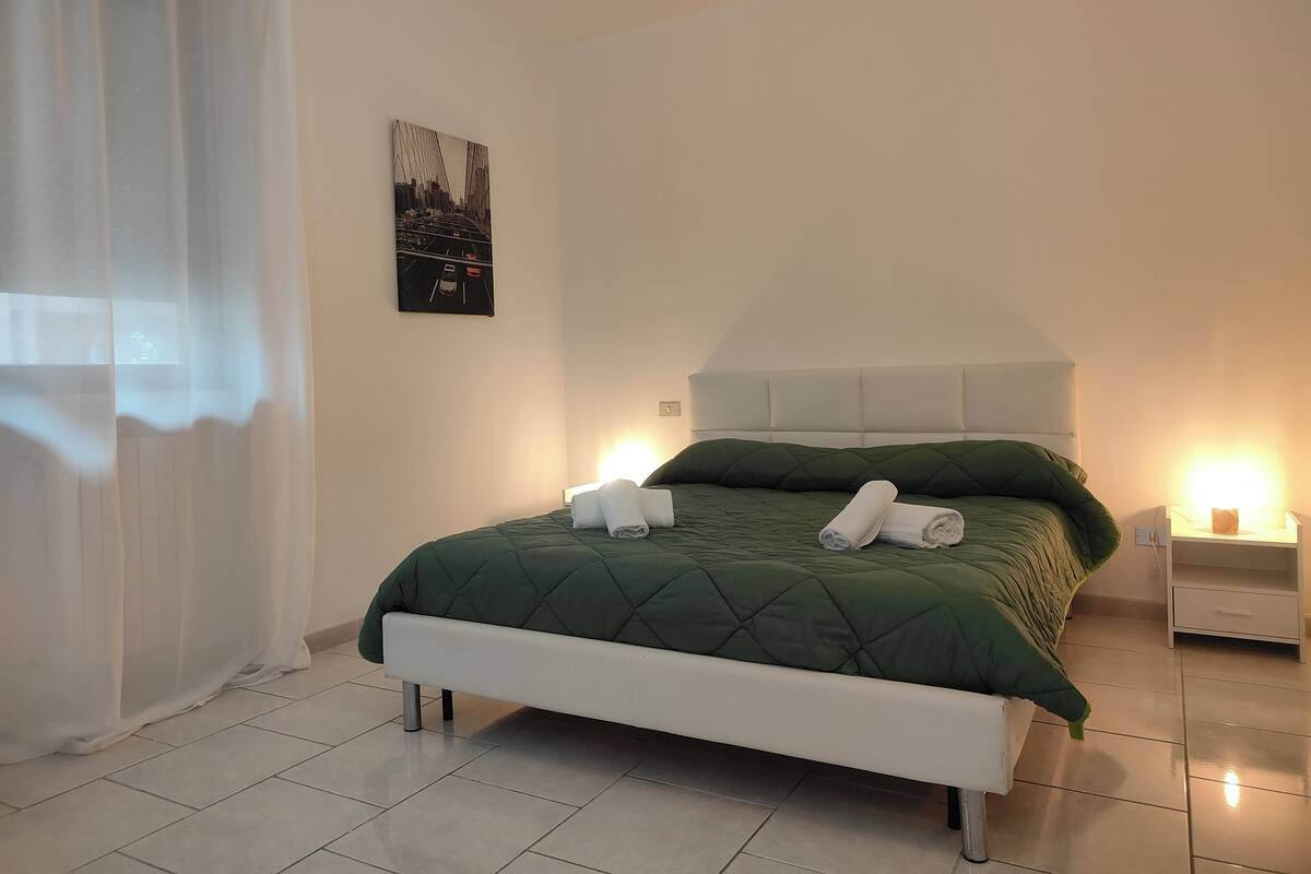 Una cama bien hecha con colcha verde y dos toallas enrolladas, cabecera blanca y mesita de noche con lámpara.