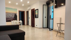 A modern living room with a sofa, a dining table with chairs, a TV mounted on the wall, and a painting of a person meditating. - CconfortHotels eXtramurale Apartment (Bari)