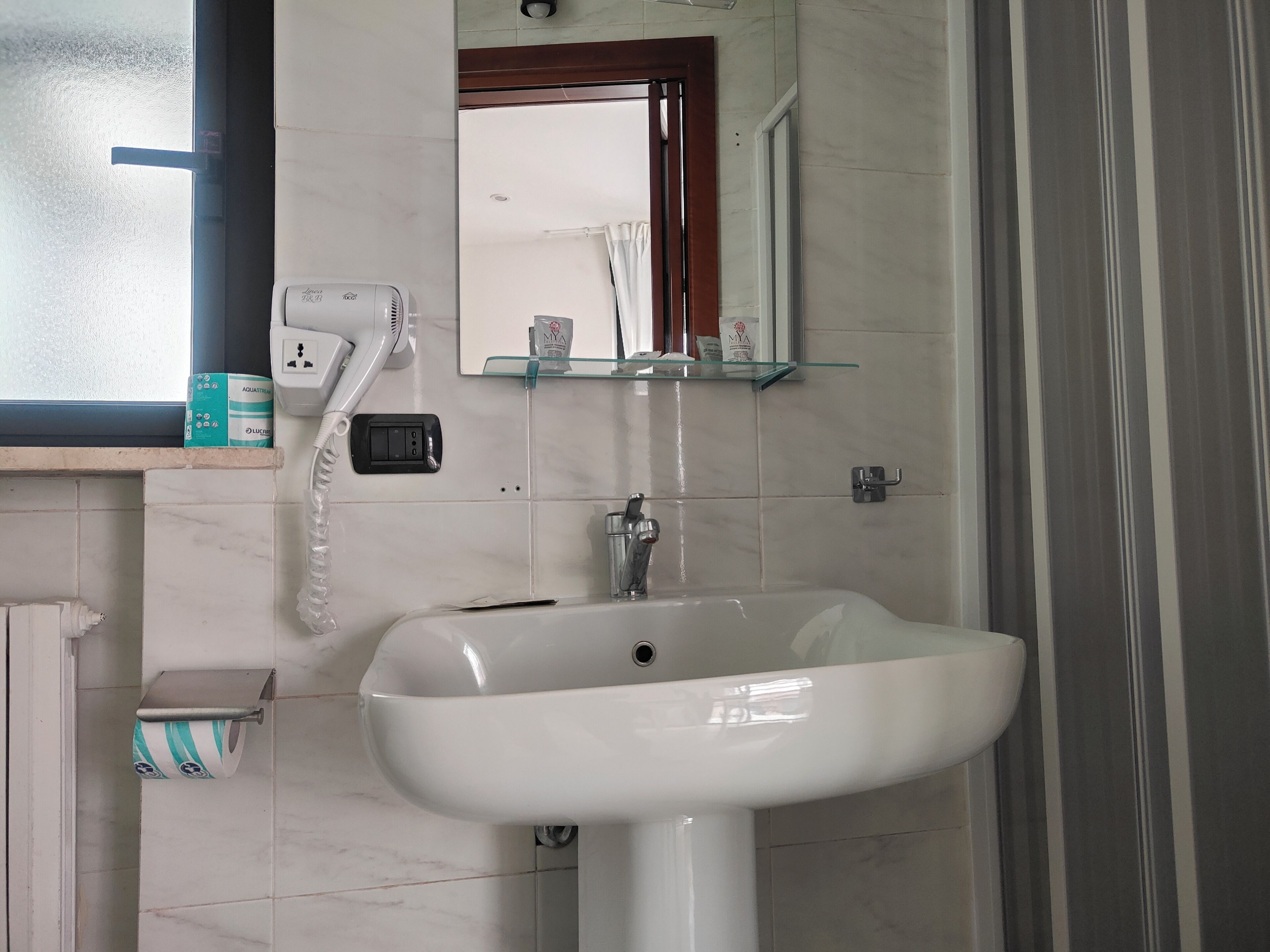A bathroom with a white sink, a mirror, a glass shelf, a hairdryer, and a toilet paper holder.