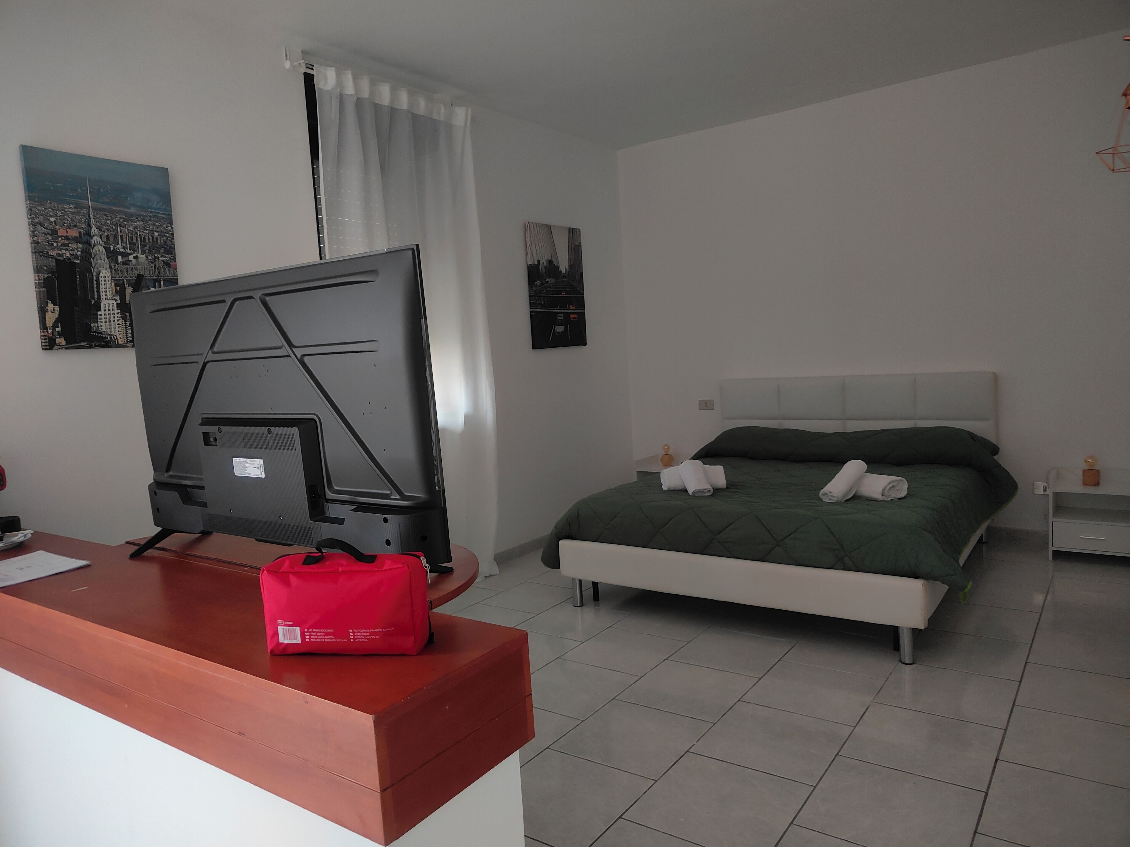 A hotel room with a bed, a desk with a television, and a red bag on the desk.