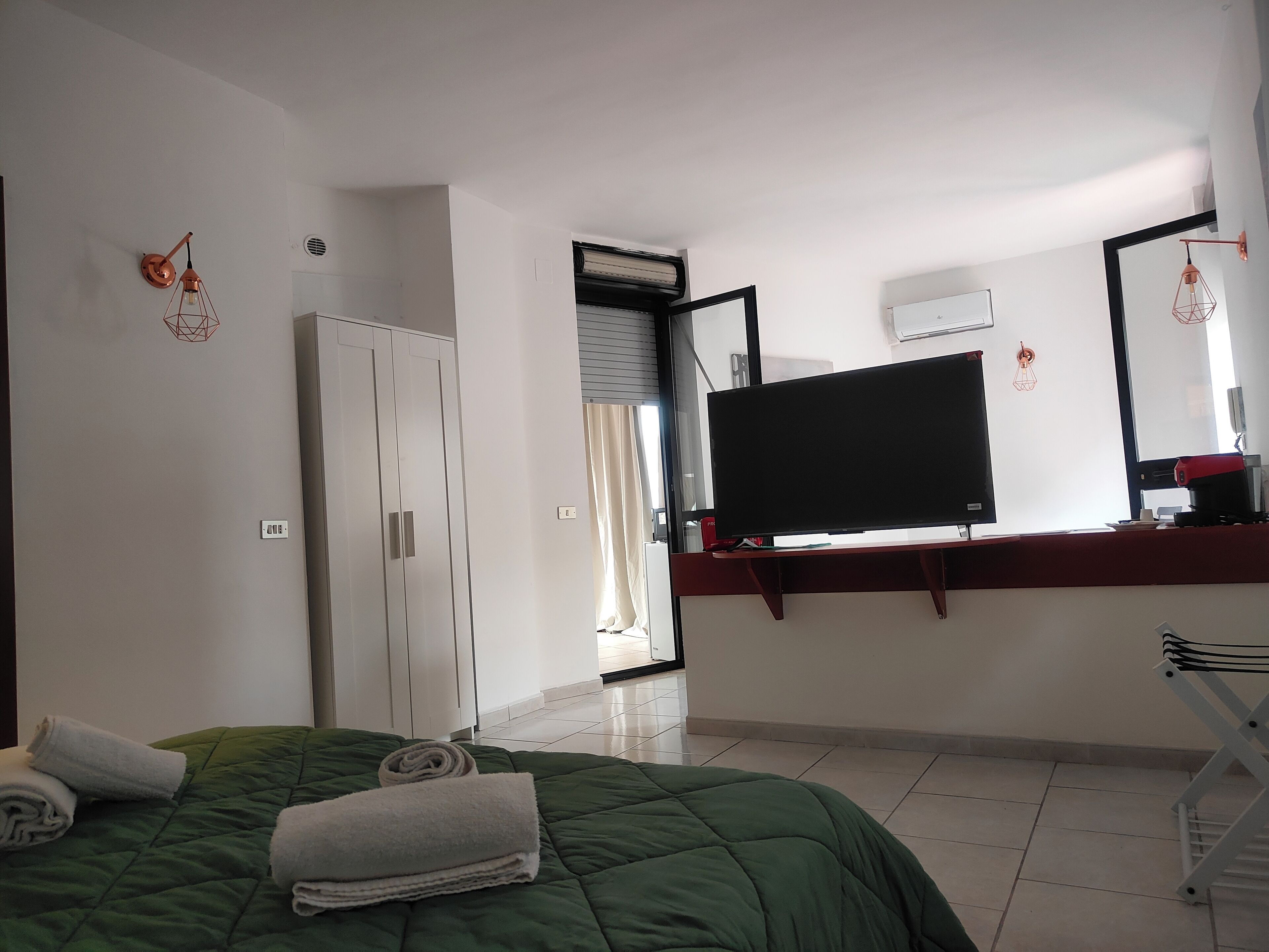 Un dormitorio con una cama verde, un televisor de pantalla plana montado en la pared, un armario y una ventana con cortinas.