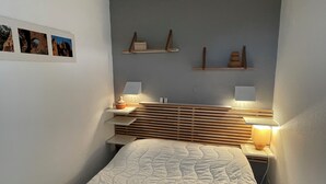A bedroom with a bed, two bedside tables with lamps, a wooden headboard, and a wall-mounted shelf with decorative items. - T1 with exceptional sea view-HAMEAU CAP ESTEREL- VILLAGE VACANCES -AGAY (Saint-Raphaël)