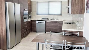 Une cuisine moderne équipée d’un réfrigérateur, d’un four, d’un four à micro-ondes et d’un lave-vaisselle. On y trouve une table à manger avec des chaises et une fenêtre avec des stores.