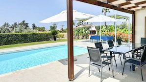 Une piscine avec un patio couvert, des sièges extérieurs et une vue sur un jardin et une maison en arrière-plan.