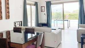Un salon moderne avec une table à manger en verre, des canapés blancs et une vue sur une piscine et la verdure environnante.
