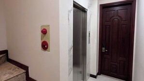 Un pasillo con pared de mármol, un botón de emergencia rojo, un ascensor de mármol y una puerta de madera oscura.