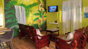 Quarto com um mural de uma cachoeira e folhagem, mobiliário de madeira, uma televisão e uma janela com cortinas.