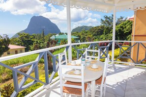 A covered outdoor seating area with a table set for two, overlooking a lush garden and a mountain. - 1-bedroom condo in charming Choiseul with AC, WiFi. Book now (Castries)