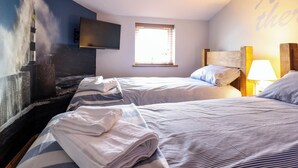 A hotel room with two beds, a TV mounted on the wall, a window with blinds, and a wall with a partial view of a lighthouse.