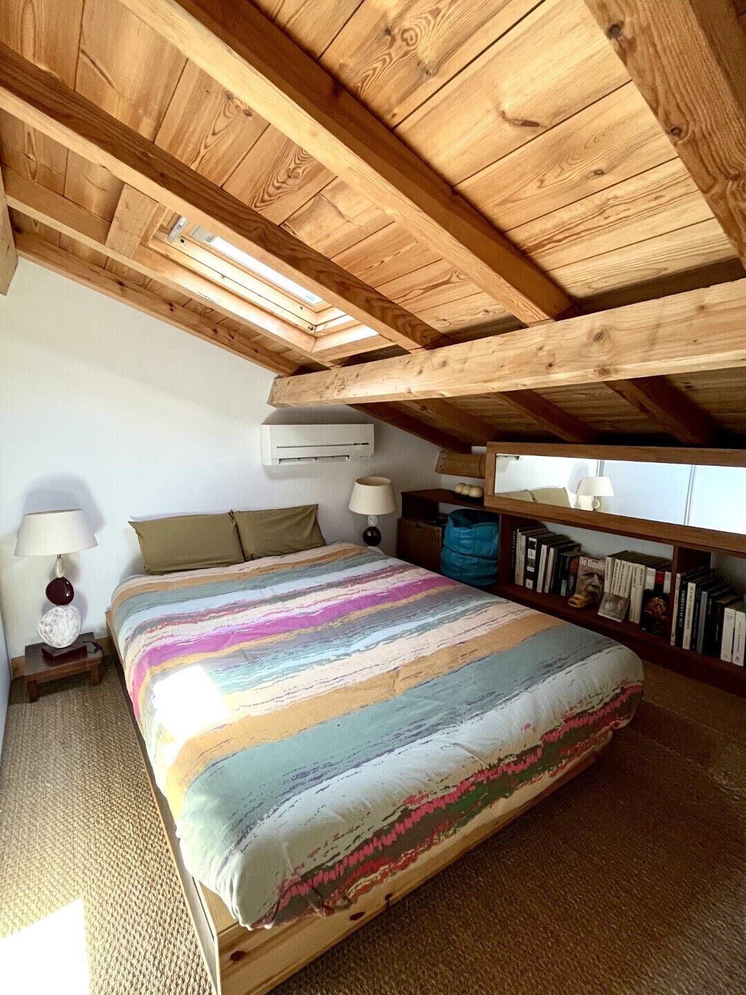 Ein Schlafzimmer mit geneigter Decke, einem Bett mit farbenfroher Streifenauflage, einer Nachttischlampe, einem Nachttisch, einem Bücherregal und einer Klimaanlage.