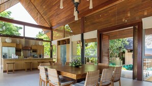 Un comedor amplio con una mesa de madera y sillas de mimbre, ventanas grandes que ofrecen vistas a la vegetación, y una cocina moderna con electrodomésticos de acero inoxidable.