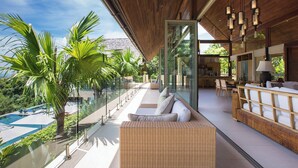 Un área de descanso moderna al aire libre con balaustrada de vidrio, mobiliario de mimbre y vistas a una piscina y vegetación.