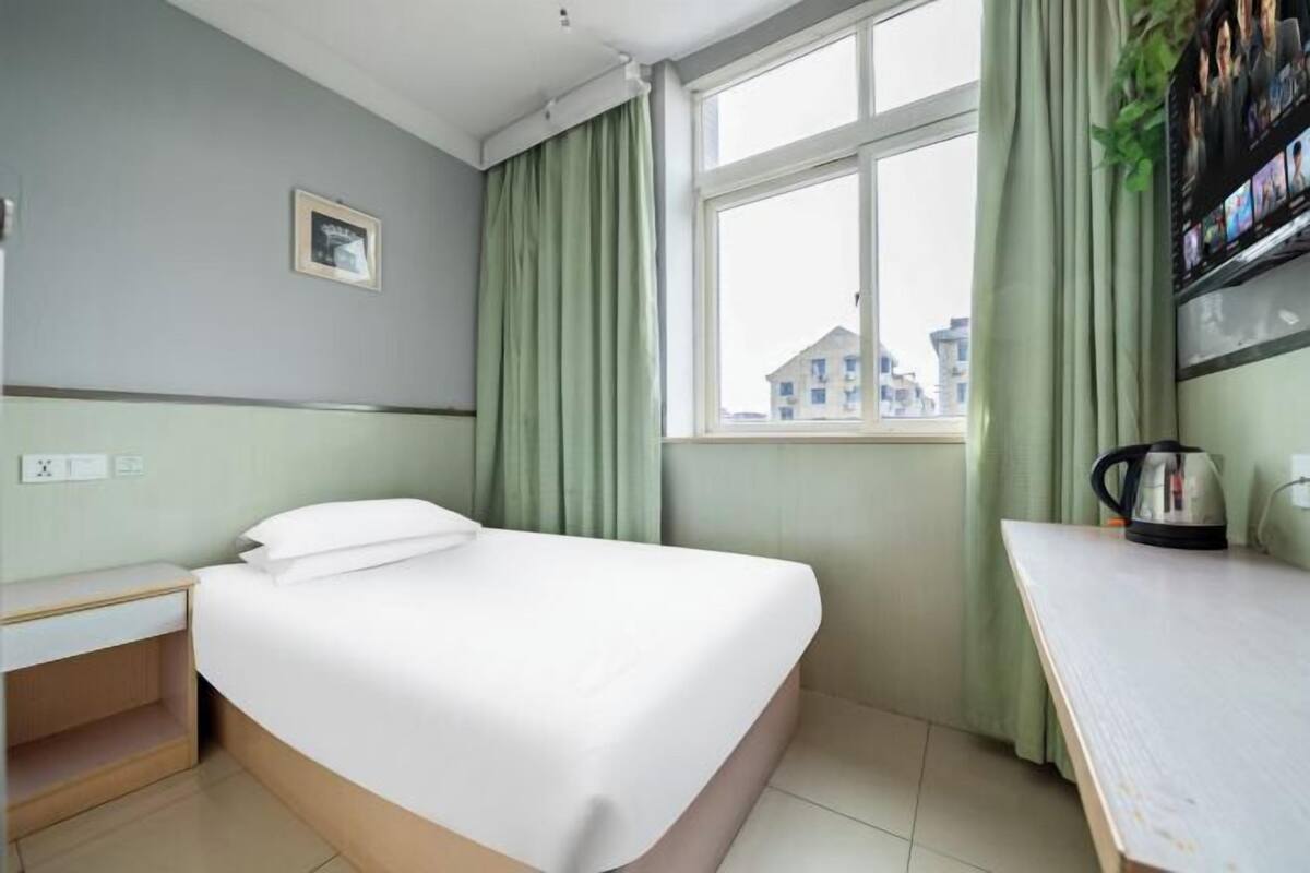 A hotel room with a bed, a desk, a window with curtains, and a view of buildings outside.