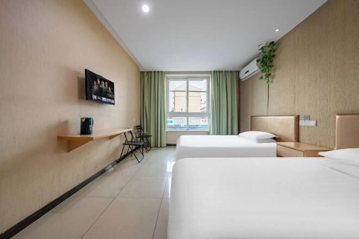 A hotel room with two beds, a TV on the wall, a desk with chairs, and a window with curtains.