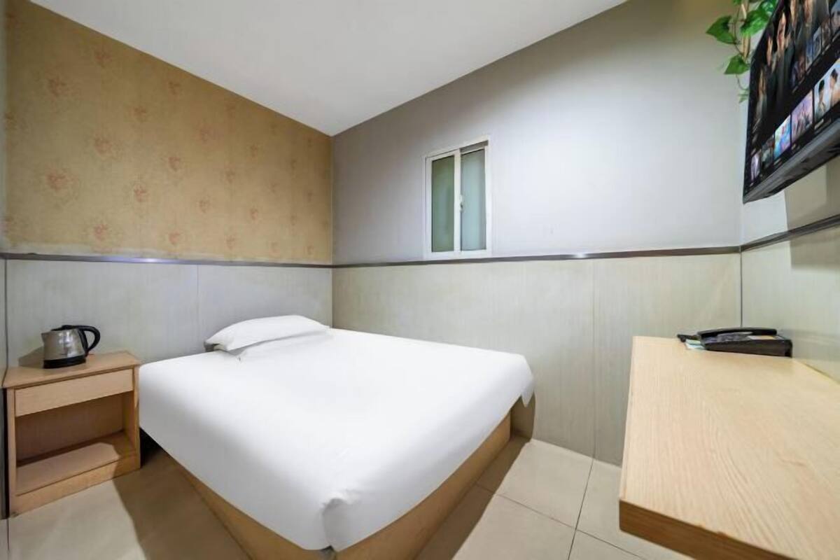A hotel room with a bed, a small bedside table with a kettle, a window, and a TV mounted on the wall.