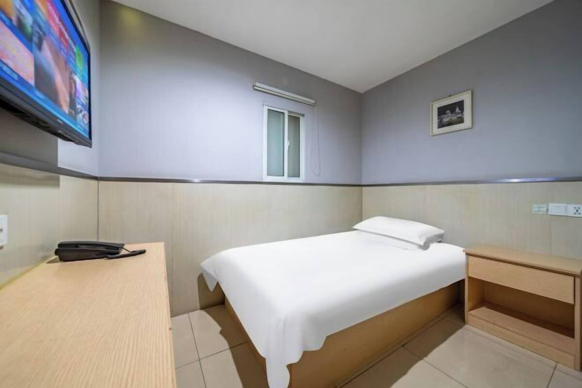 A hotel room with a bed, bedside table, a desk, a TV, a window, and a picture on the wall.