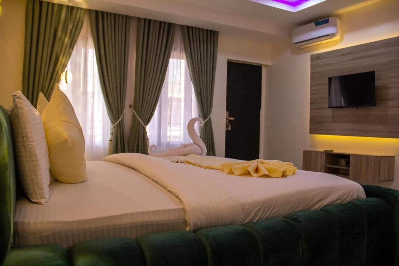 A hotel room with a large bed, green upholstered headboard, a flat-screen TV mounted on the wall, and a built-in air conditioning unit.