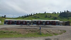 Eine Reihe kleiner, zweistöckiger Hütten in Rot- und Beigetönen, vor einer sanften Hügellandschaft mit vereinzelten Bäumen.
