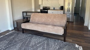 Ein Wohnzimmer mit einer Couch, einem Couchtisch und einem Teppich. Im Hintergrund ist eine Küche mit Schränken und Geräten zu sehen.
