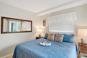 A bedroom with a large bed, a mirror, a window with blinds, and a nightstand with a lamp. - Beachfront  - Fantastic Beach Vacation for Family Fun (Gulf Shores)