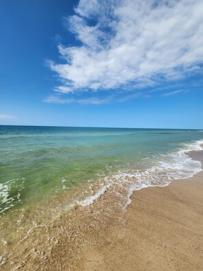 A beach with clear water, a sandy shore, and a blue sky with clouds.