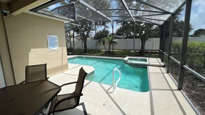A covered outdoor pool area with a table and chairs, a hot tub, and a view of the surrounding trees and fence.