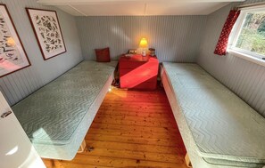 Et lille, hyggeligt værelse med to senge, trægulv, en rød kommode, en lampe og billeder i rammer på væggene.
