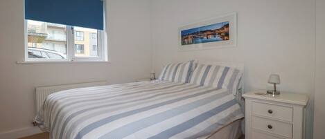 A bedroom with a bed, a nightstand, a lamp, a window with a view of buildings, and a framed picture on the wall.