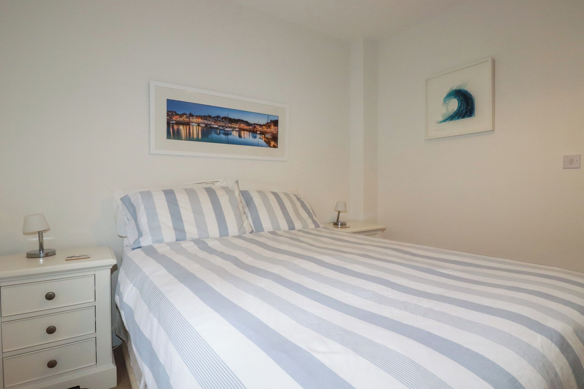 A bedroom with a bed, two bedside tables, a nightstand, a framed picture of a coastal scene, and a framed abstract painting.