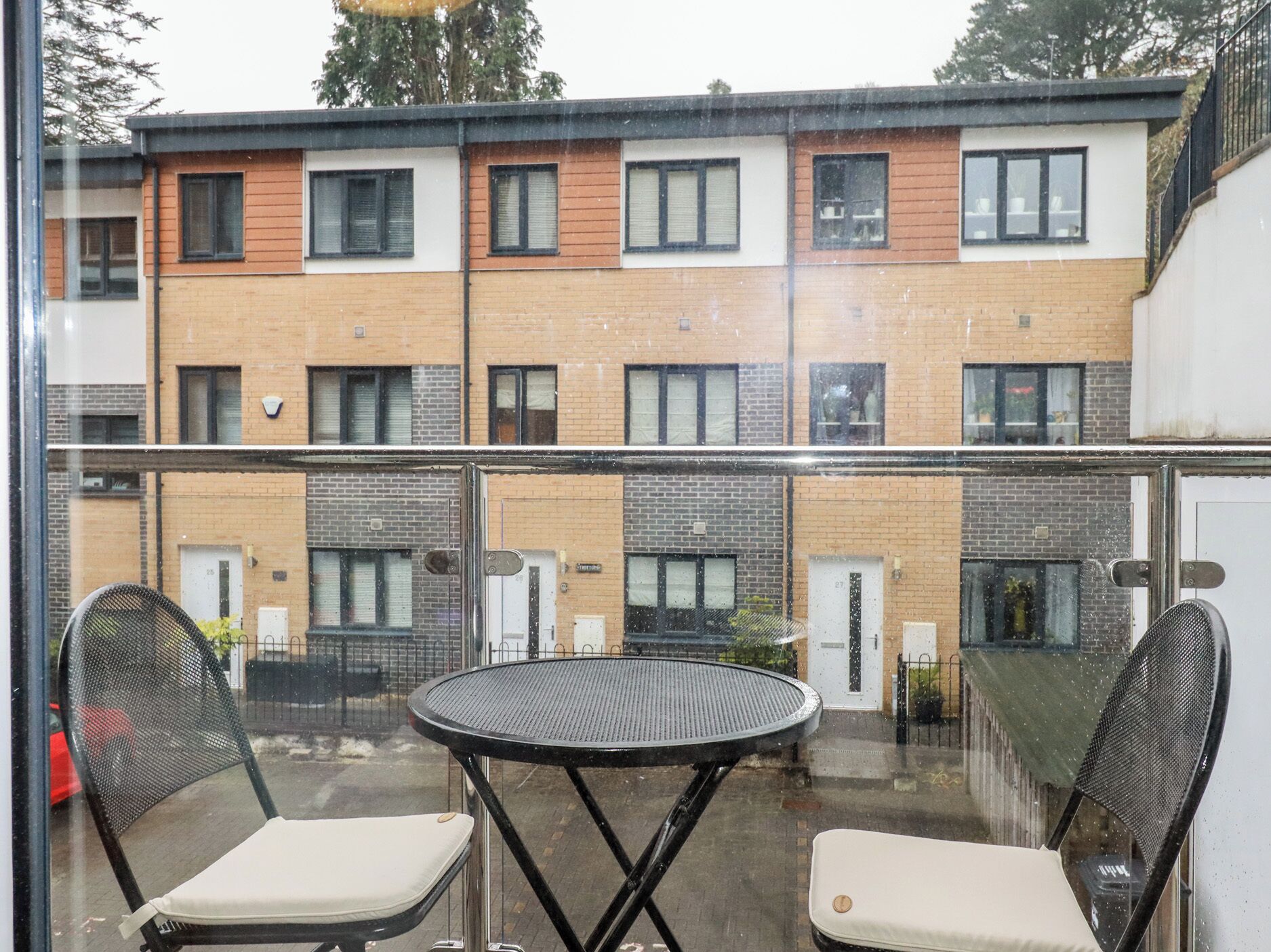 Een balkon met een ronde tafel en twee stoelen, met uitzicht op een rij bakstenen gebouwen met meerdere ramen.