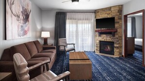 Habitación de hotel con chimenea de piedra, un sofá, una silla, una mesa de centro y un televisor montado en la pared.