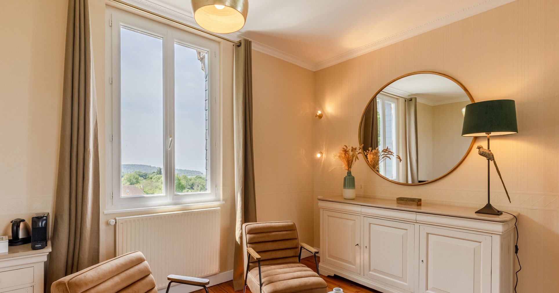 Een kamer met een groot raam, een stoel, een tafeltje, een spiegel en een lamp.