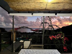 Une terrasse offrant une vue sur la mer et le coucher de soleil, comprenant une table et des chaises, une terrasse en bois et une pergola ornée de fleurs.